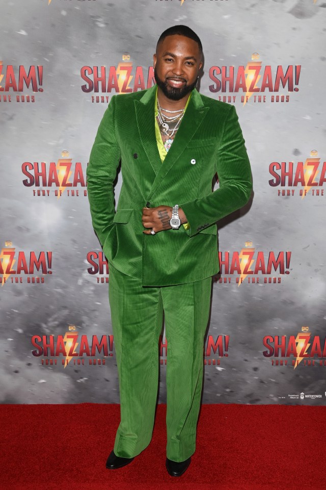 a man in a green suit stands in front of a poster for shazam