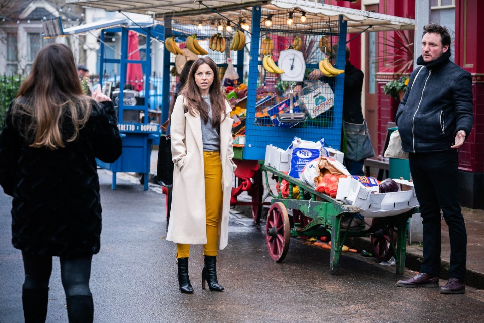 a woman taking a picture of a man standing in front of a fruit stand that says ' metro ' on it