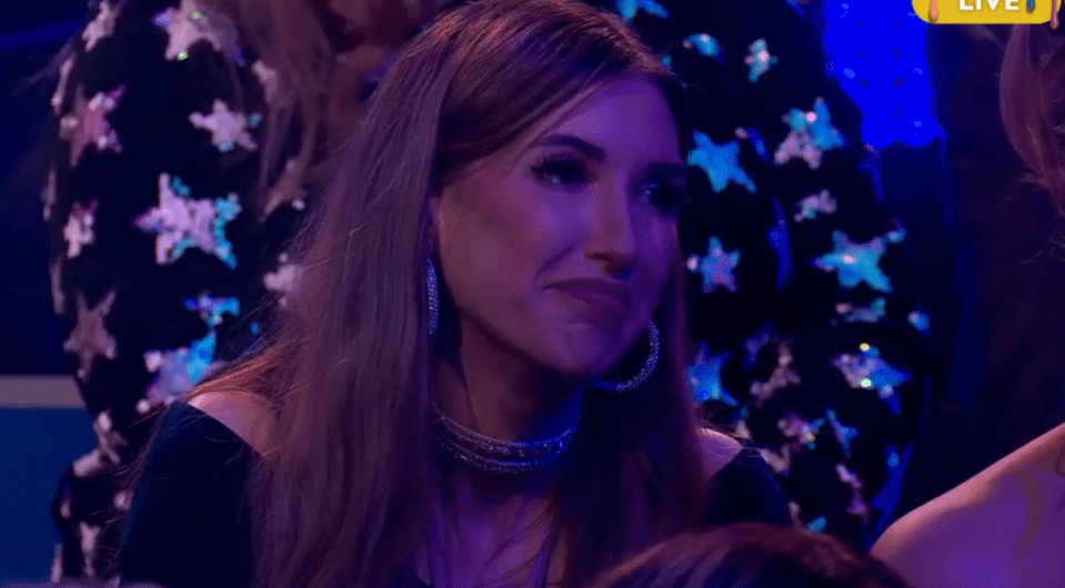 a woman wearing hoop earrings and a choker is sitting in front of a live screen