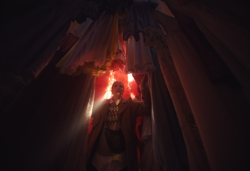 a man in a mask stands in a dark room with clothes hanging from the ceiling