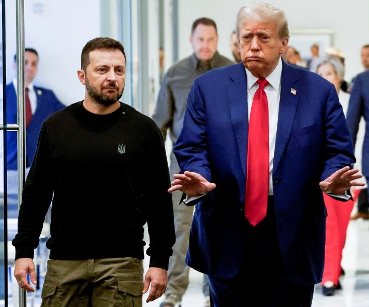 a man in a black sweater with a eagle on it walks next to a man in a blue suit and red tie