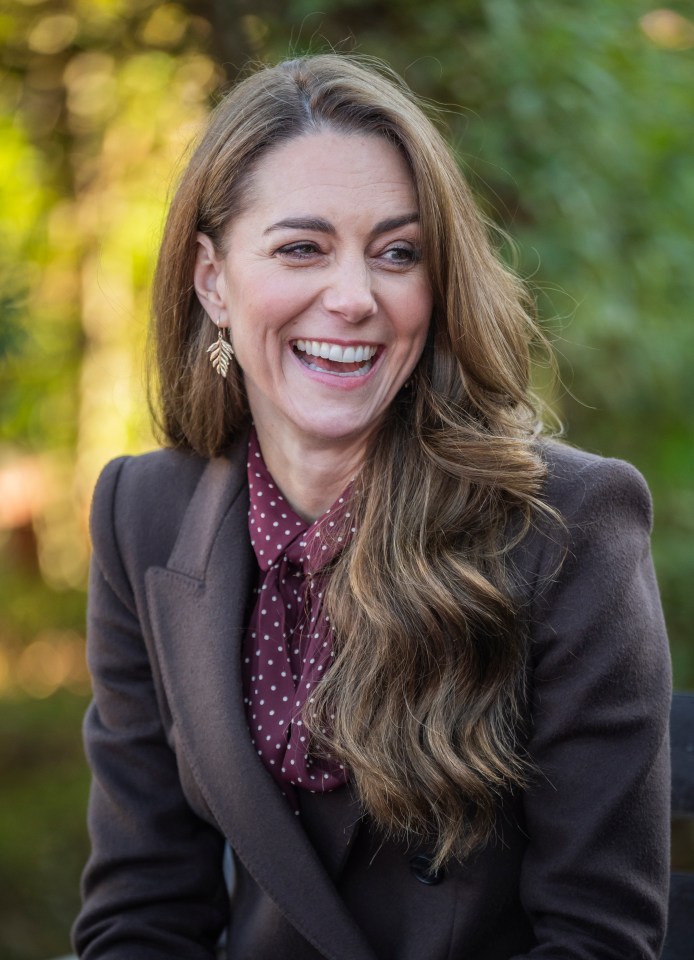 a woman wearing a polka dot shirt and a brown coat is smiling