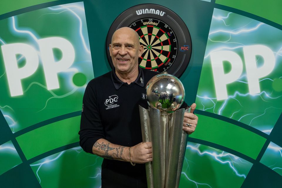 a man holding a trophy in front of a winmau dart board