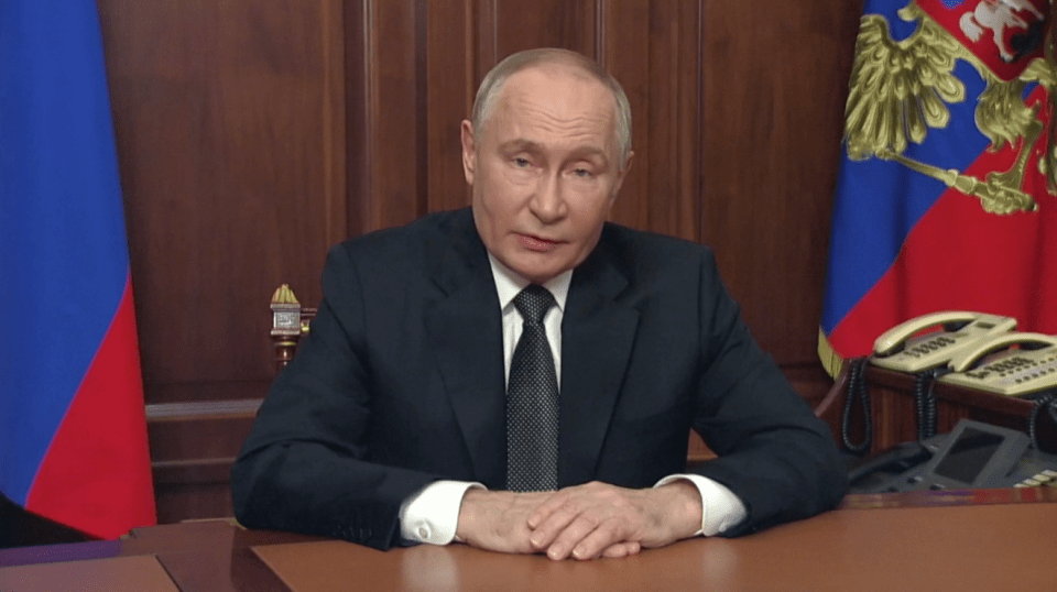 a man in a suit and tie sits at a desk with his hands folded