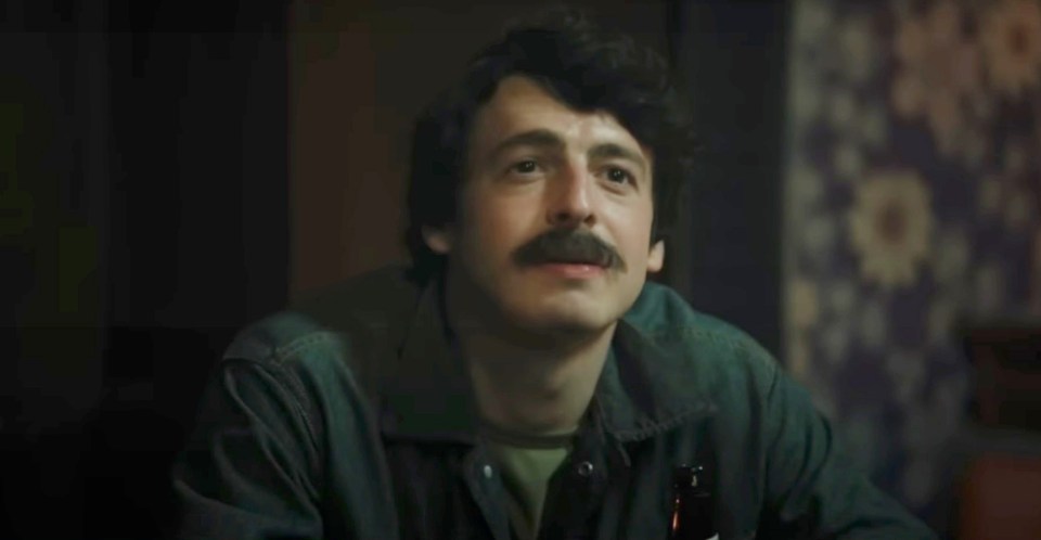 a man with a mustache is sitting at a table holding a bottle of beer .