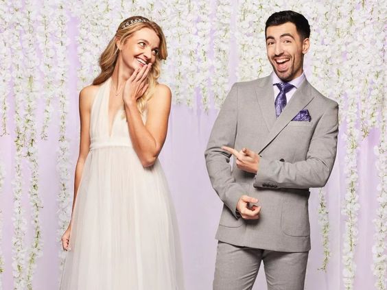 a bride and groom are posing for a picture in front of a purple background with white flowers .