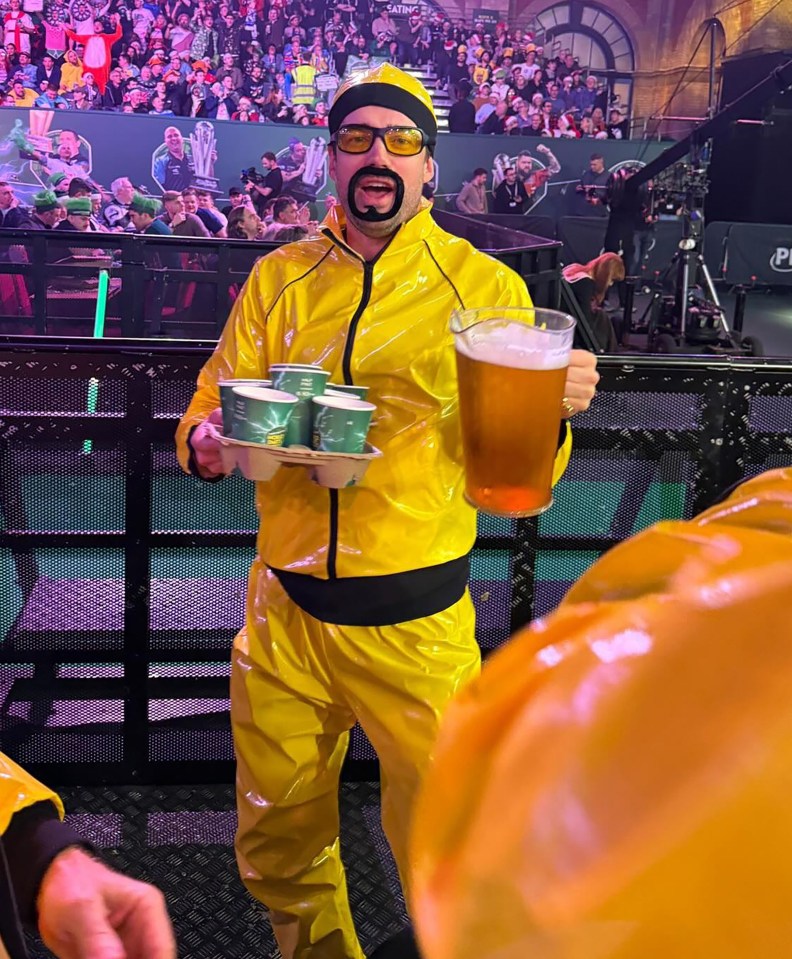 Jack Whitehall in a yellow jumpsuit at a darts championship, holding a beer and drinks.