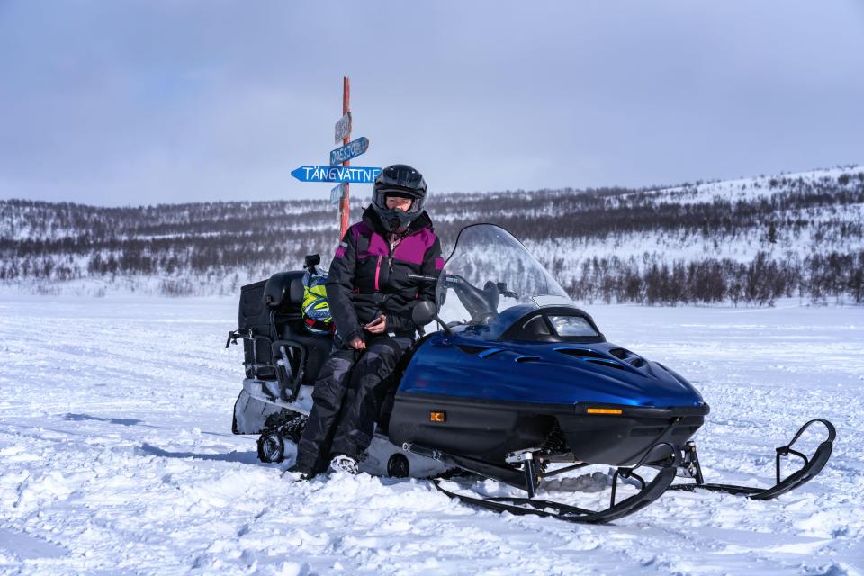 A middle aged caucasian woman wearing a helmet and winter clothing sitting on a snowmobile in Lapland mountains