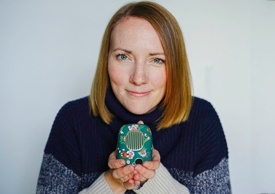 A woman holding a hot water bottle and a power bank in her hands, she is wearing a red top and jeans and is sitting on a sofa.
