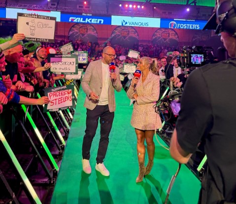 Two reporters interviewing fans on a green carpet.