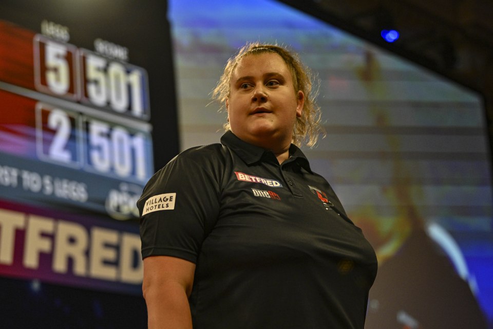 A woman with long blonde hair, wearing a blue top, white skirt, and black shoes, is standing on a stage with a microphone in her hand. There is a dart board in the background, and the words "Winter Gardens Blackpool" can be seen on a banner.