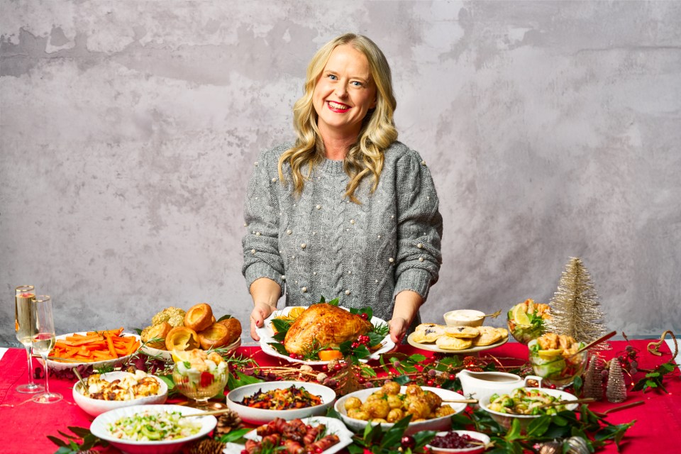 A woman presents a Christmas dinner with roasted turkey and various side dishes.