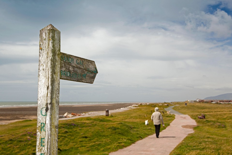 Wooden signpost showing directions to Sandy Gap and Bridleway, with a person walking a dog on a coastal path.