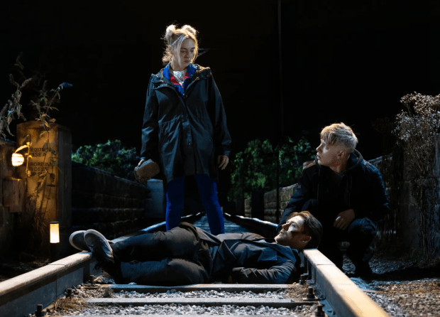 Coronation Street scene: a man lies on train tracks as two young people look on.