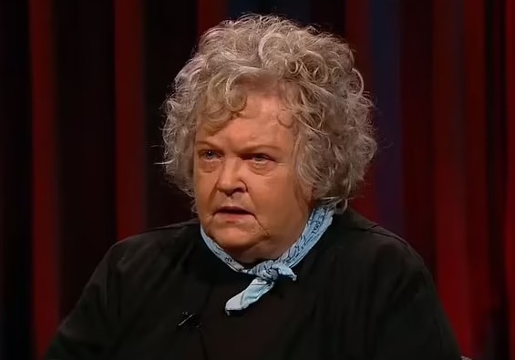 A woman with curly gray hair looks thoughtfully at the camera.