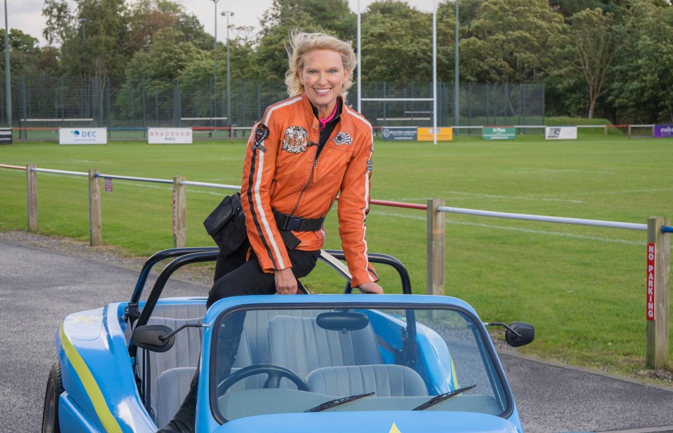 Anneka Rice in an orange jacket sits on a light blue car.