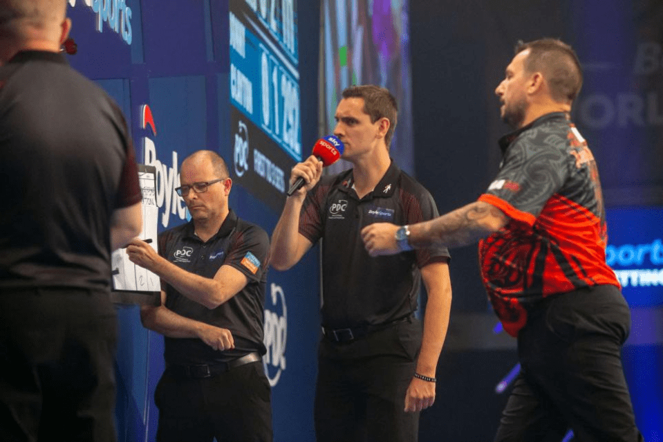 Three men at a darts match; one is being interviewed, one is recording the score, and one is a player.