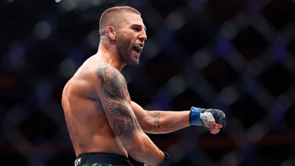 A male MMA fighter in a cage, mouthguard in, gesturing.