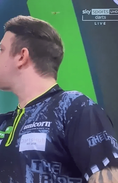 Close-up of a darts player's back, wearing a dark shirt with "Unicorn" and "The Rise" logos.  Sky Sports UHD Darts Live is visible in the background.