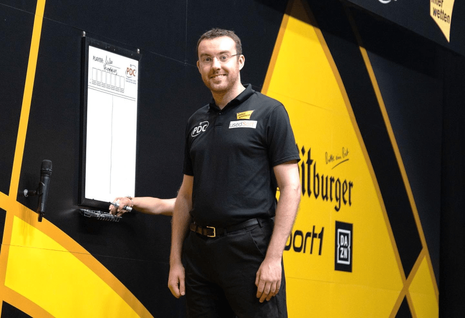 A man stands in front of a dartboard scoreboard.