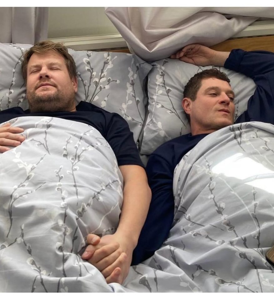 Behind-the-scenes photo of James Corden and another man in bed holding hands.