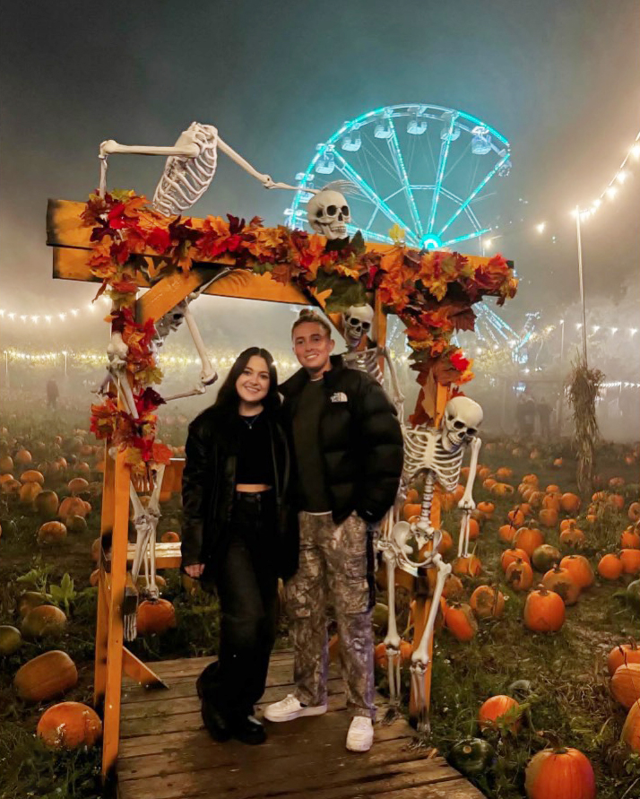 Couple posing at a pumpkin patch with skeletons and a Ferris wheel in the background.