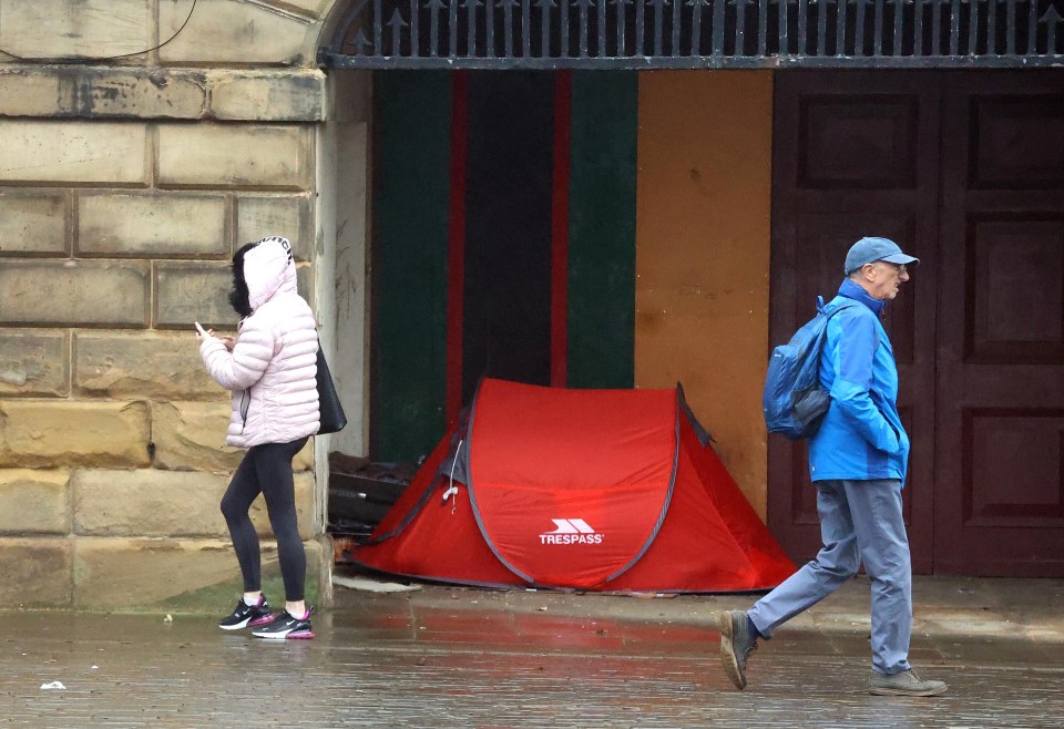 A red pop-up tent sits on the sidewalk near a person using a phone and a person walking by.