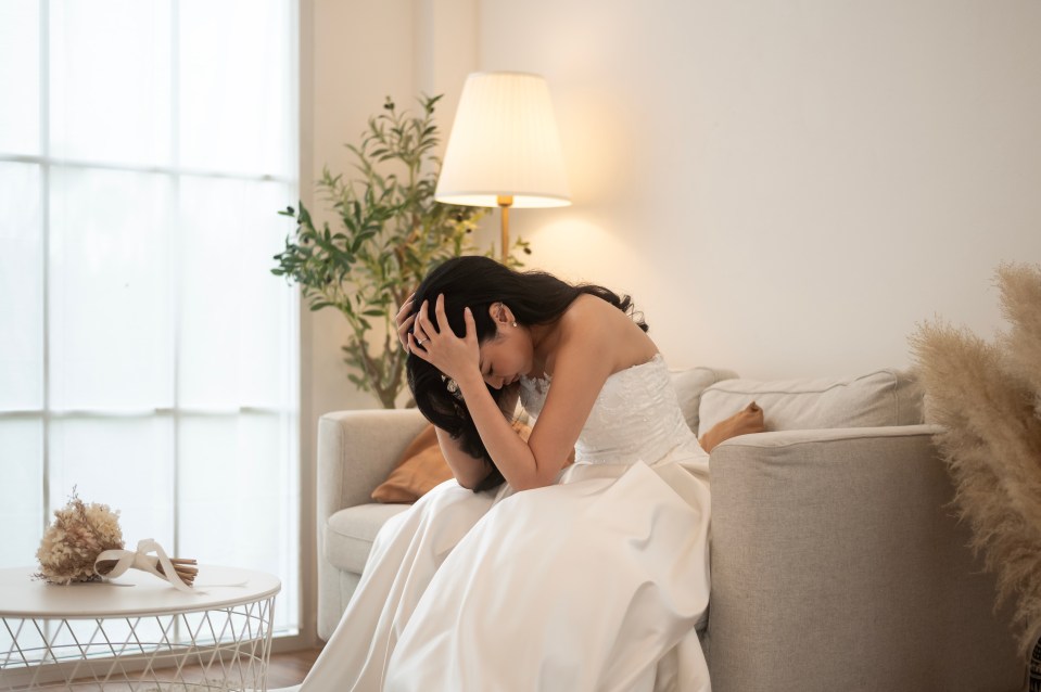 A bride in a wedding dress sits on a couch, crying and holding her head in her hands.