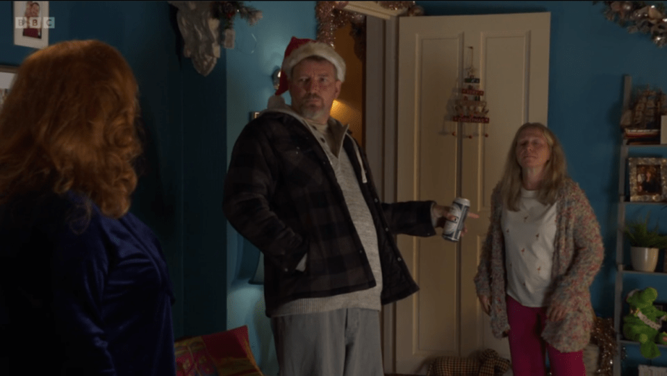 A scene from EastEnders showing a man in a Santa hat holding a beer can, with two women looking on.