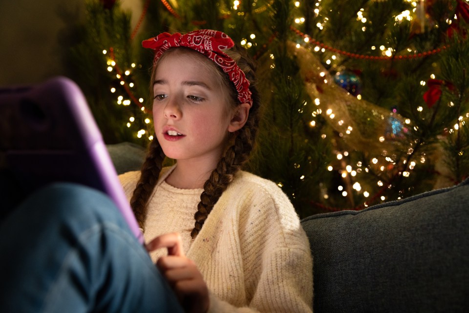 A close-up shot of a young girl sitting comfortably on a sofa in the living room of her home in South Shields, North East England. She is playing on a digital tablet, her face illuminated by the screen. In the background there is a twinkling Christmas tree visible. She has a neutral expression, braided hair and is wearing a headband.</p>
<p>Video similar to this scenario available.