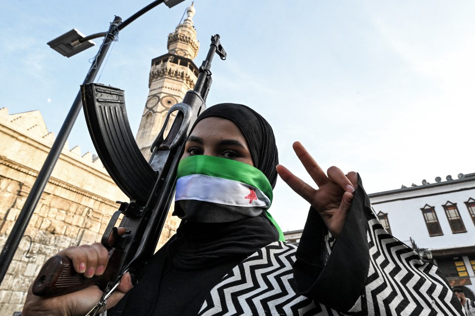 Woman in Damascus holding an assault rifle, her face masked by the Syrian opposition flag, makes a peace sign.