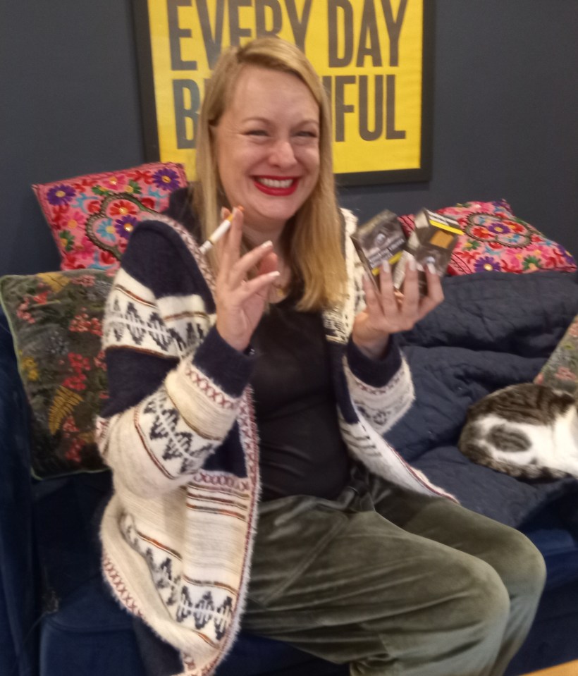 Woman sitting on a couch holding cigarettes and a cat sitting beside her.
