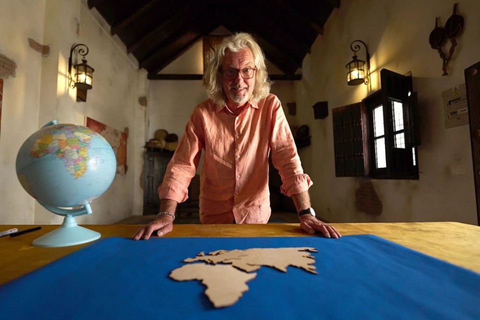 James May leaning on a table with a globe and a world map.