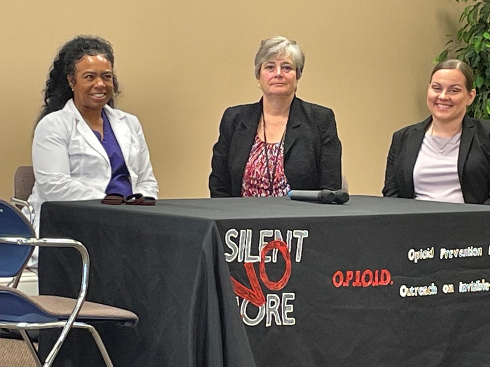 Three women sit at a table with a "Silent No More" opioid prevention banner.
