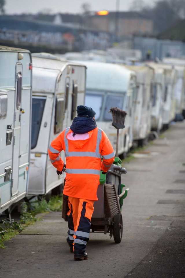A sanitation worker in an orange uniform pushes a cleaning cart past a row of caravans.