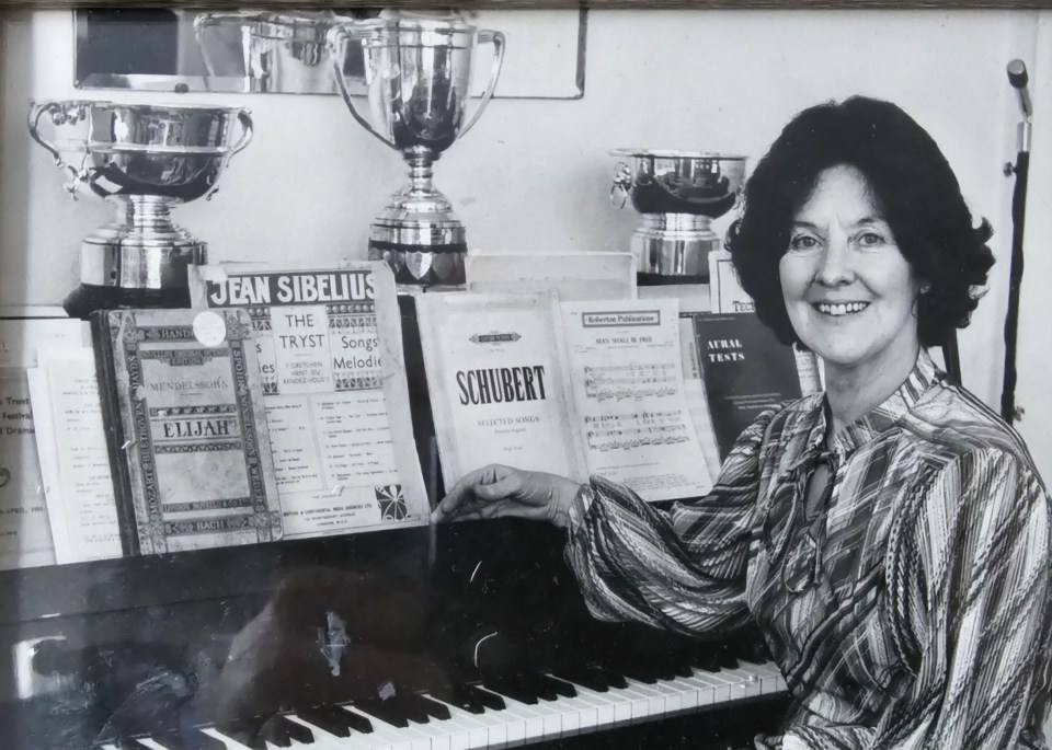 Black and white photo of a woman sitting at a piano with sheet music and trophies.
