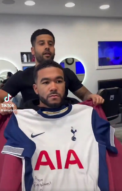 Man getting a haircut in a barbershop while wearing a signed Tottenham Hotspur jersey.