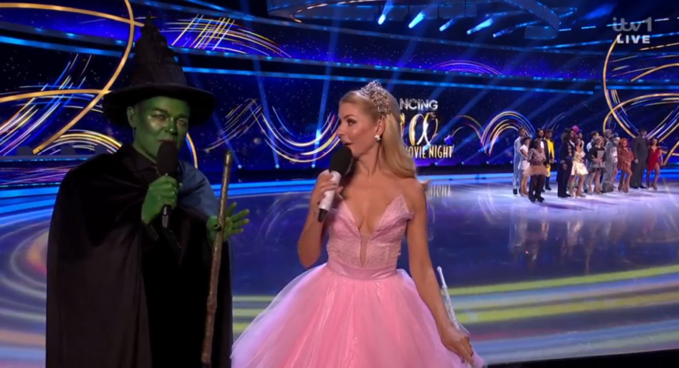 Screenshot of a woman in a pink dress interviewing a person dressed as the Wicked Witch of the West on a skating rink.