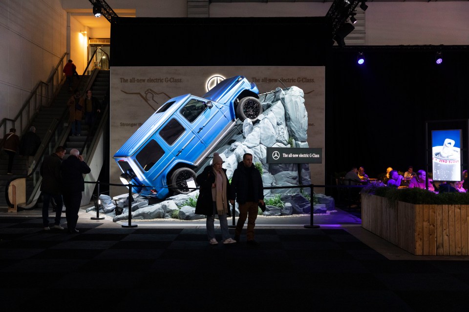 A blue electric G-Class SUV displayed at a car show, appearing to climb a rock formation.