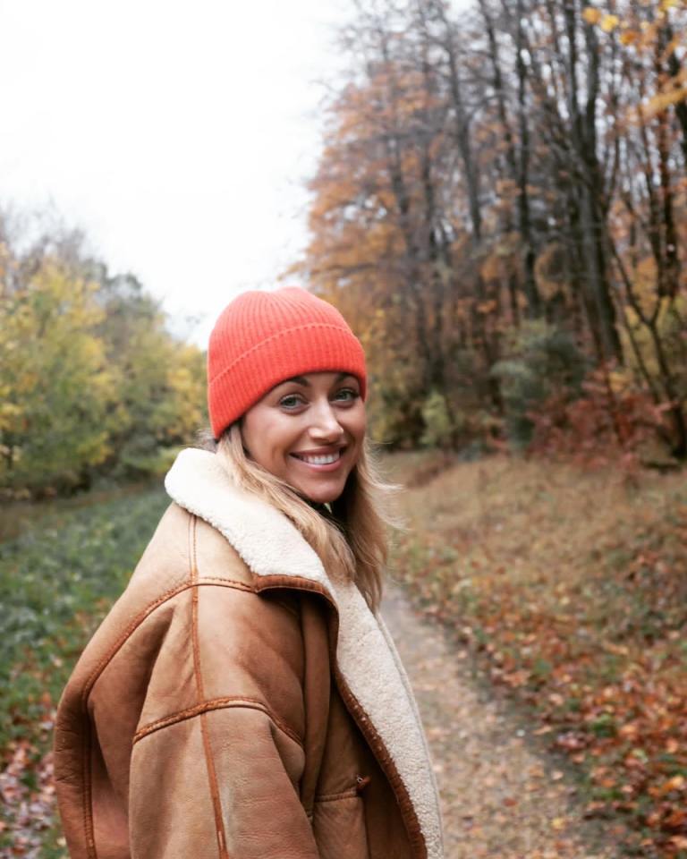 Woman in orange beanie and brown jacket smiles in autumn woods.