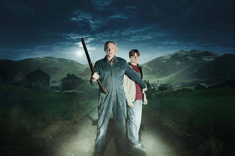 Two men stand on a road at night, one holding a shotgun, with a rural landscape behind them.