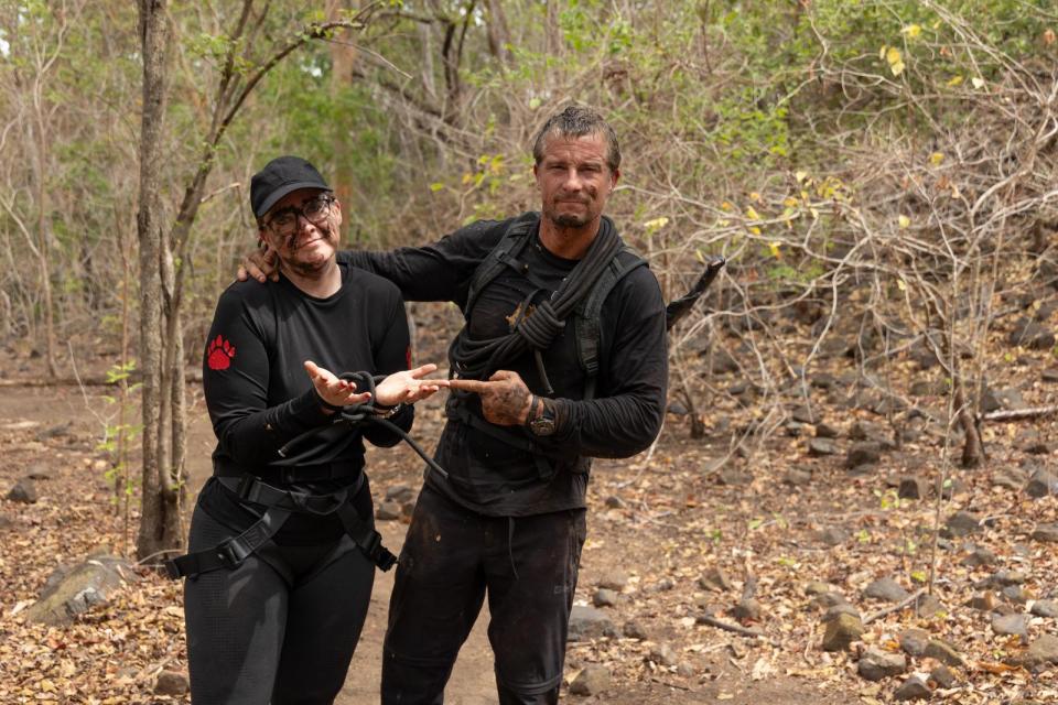 Katie Begley and Bear Grylls in a forest, her hands bound with rope.