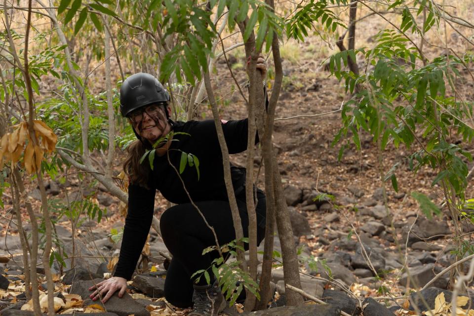 Woman in helmet and camouflage crouching in bushes.