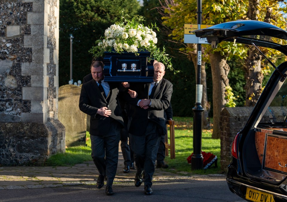 Pallbearers carrying a coffin draped with white flowers at a funeral.