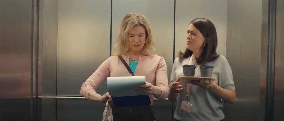 Two women in an elevator; one reviews paperwork, the other carries coffee.