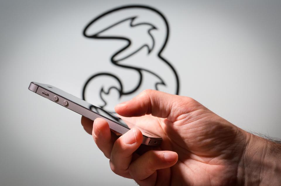 A hand holding a mobile phone in front of a Three Mobile network logo.