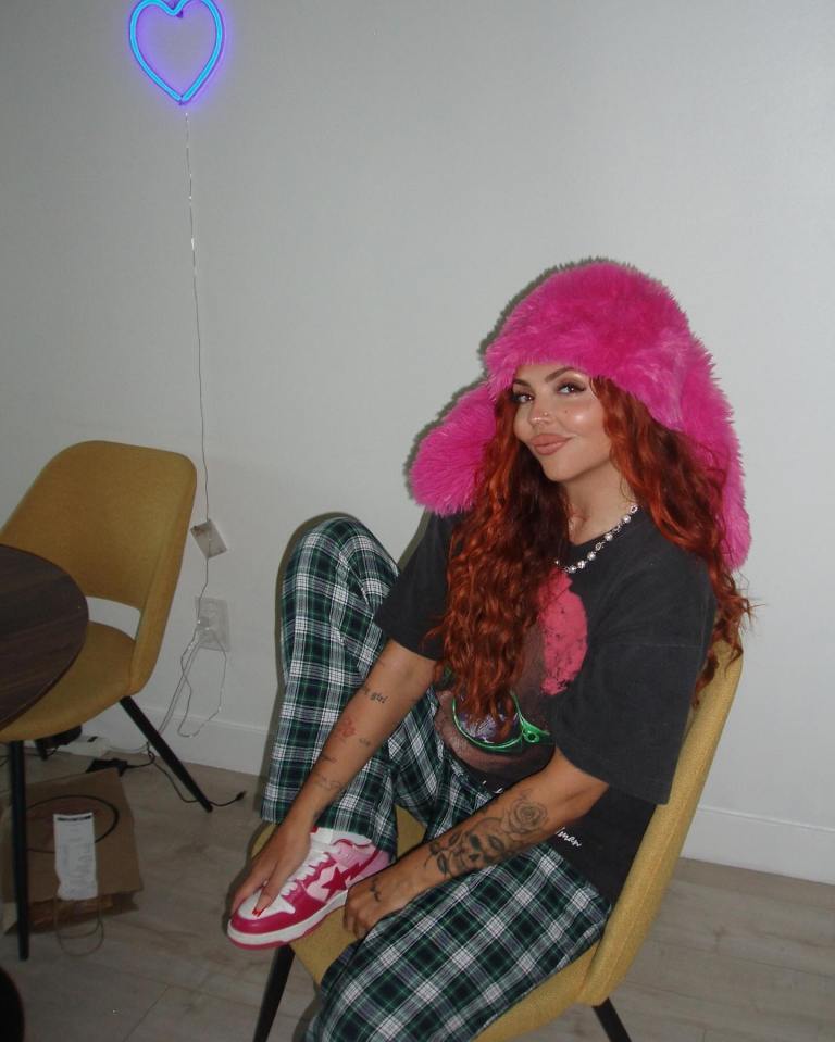 Jesy Nelson sitting in a chair wearing a pink hat and plaid pants.