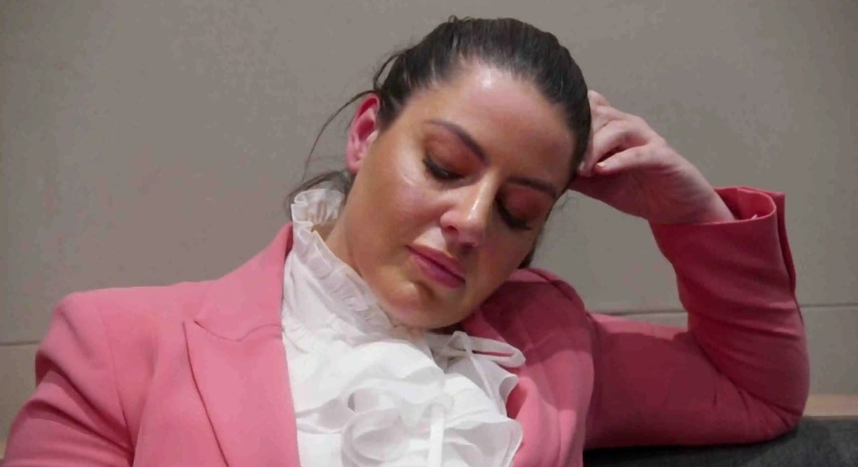 A woman in a pink blazer and white ruffled blouse sits with her hand to her head.