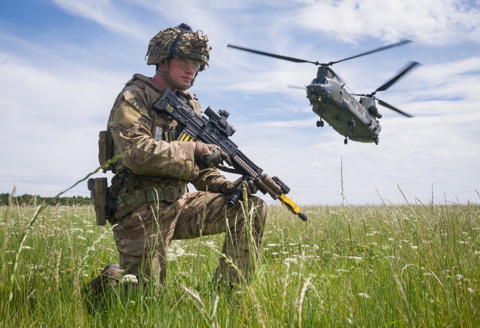 British soldier kneeling in a field with a helicopter overhead.