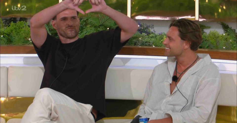 Two men sitting on a couch, one making a heart shape with his hands.